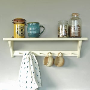 May include: A white shelf with hooks underneath. The shelf has a blue and green ceramic jug, a yellow and brown coffee jar, and two glass jars with lids. There are two brown ceramic mugs hanging from the hooks and a white tea towel with a blue pattern hanging from the hooks.
