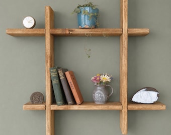 May include: A rustic, wooden wall shelf with a cross-shaped design. The shelves display a small clock, a blue ceramic pot with a plant, books, a vase of flowers, and decorative stones. The shelf is mounted on a muted green wall.