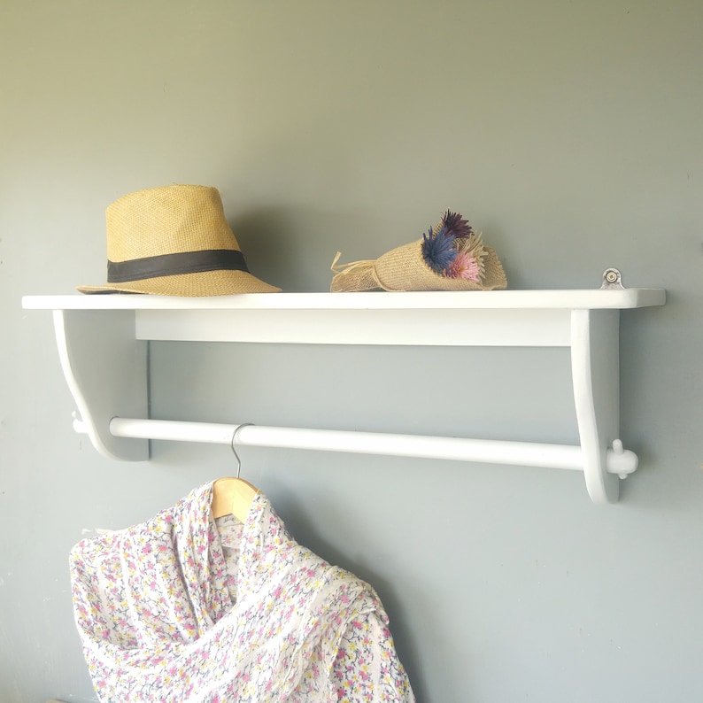 May include: White wooden shelf with a white metal rod for hanging clothes. A straw hat and a bouquet of dried flowers are on the shelf. A white and pink floral scarf is hanging from the rod.
