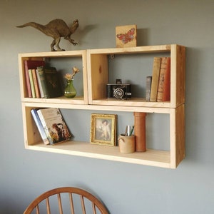 May include: A wooden shelf unit with three shelves. The top shelf features a butterfly picture and a camera. The middle shelf has books and a vase with flowers. The bottom shelf has books, a framed picture, and a pencil holder.
