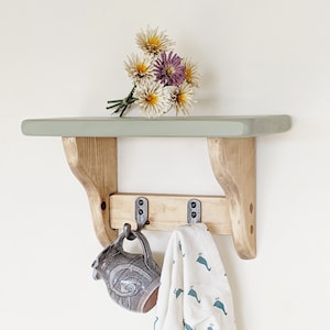 May include: A light green shelf with two hooks and a small brown ceramic jug hanging from one of the hooks. A white cloth with a blue pattern hangs from the other hook. A bouquet of yellow and purple flowers sits on the shelf.