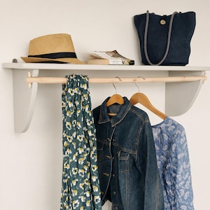 May include: A light grey wall shelf with a wooden rail and hangers, holding a straw hat, a book, a navy tote bag, a floral scarf, a denim jacket, and a blue floral top. The shelf is mounted on a white wall.