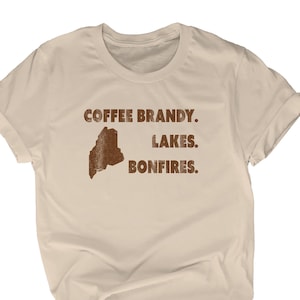 May include: A beige t-shirt with a brown outline of the state of Maine and the text "Coffee Brandy. Lakes. Bonfires."