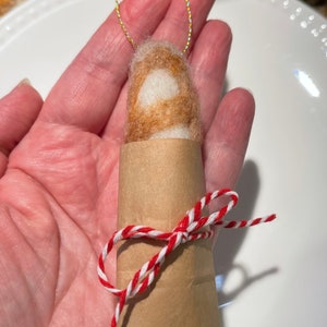 May include: A small, brown and white felted ornament wrapped in brown paper and tied with red and white twine. The ornament is hanging from a gold string.