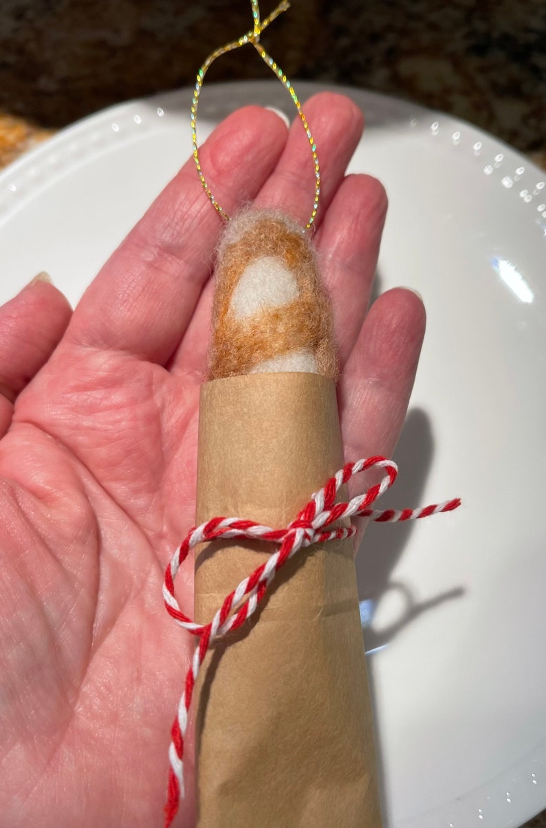 May include: A small, brown and white felted ornament wrapped in brown paper and tied with red and white twine. The ornament is hanging from a gold string.