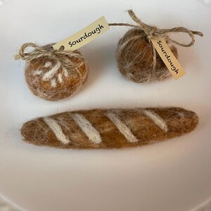 May include: Two brown felt balls with the word "Sourdough" written on small tags, and a brown felt baguette with white stripes.