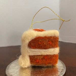 May include: A felt ornament shaped like a layered cake with orange and white frosting. The ornament has a gold string for hanging.