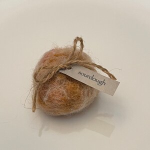 May include: A round, textured object with a light brown and orange color scheme, tied with twine and a small tag that reads "sourdough". The object is likely a decorative item or a small craft.