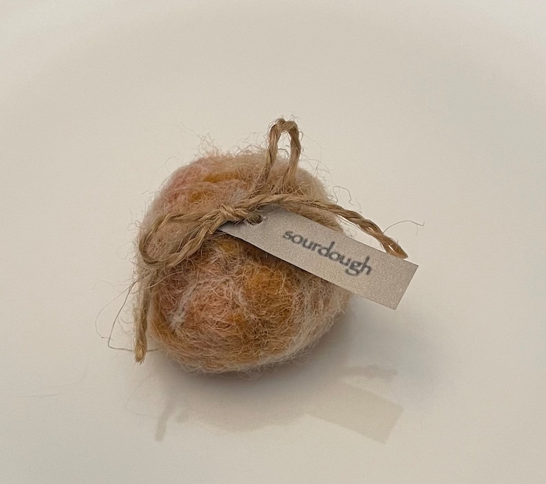 May include: A round, textured object with a light brown and orange color scheme, tied with twine and a small tag that reads "sourdough". The object is likely a decorative item or a small craft.
