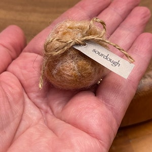 May include: A small, round, light brown object resembling a miniature loaf of bread, tied with twine and a tag that reads "sourdough". The object is held in a person's hand.