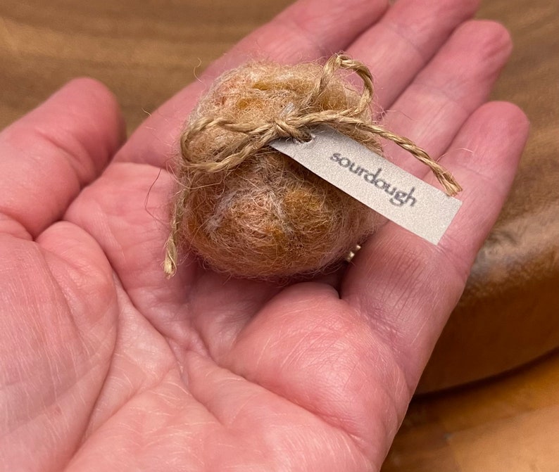 May include: A small, round, light brown object resembling a miniature loaf of bread, tied with twine and a tag that reads "sourdough". The object is held in a person's hand.