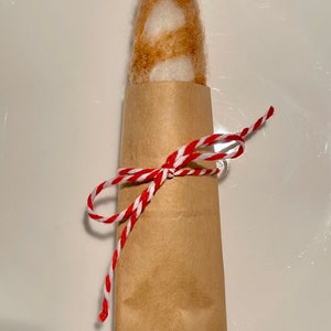 May include: A brown and white felted baguette wrapped in brown paper and tied with red and white twine.