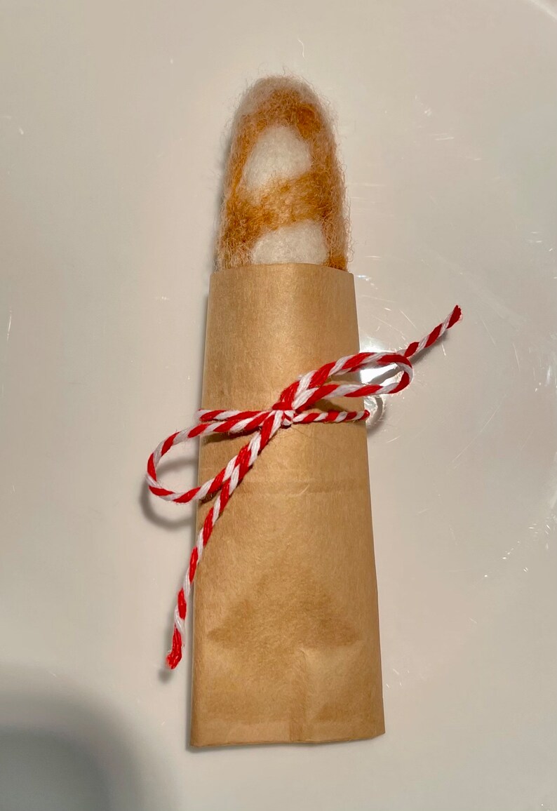 May include: A brown and white felted baguette wrapped in brown paper and tied with red and white twine.