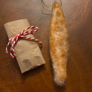 May include: A brown and white felted ornament shaped like a long, thin fish. The ornament is hanging from a gold string and is wrapped in brown paper with a red and white twine bow.