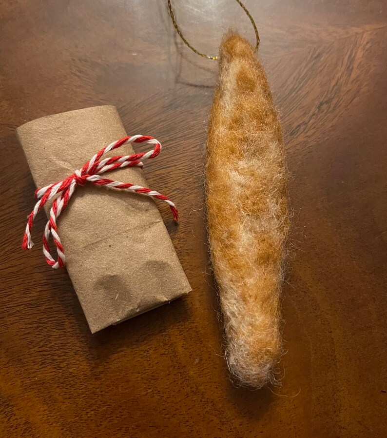 May include: A brown and white felted ornament shaped like a long, thin fish. The ornament is hanging from a gold string and is wrapped in brown paper with a red and white twine bow.