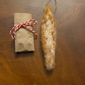May include: A brown felt ornament shaped like a baguette, hanging from a gold string. The ornament is on a brown wooden surface.