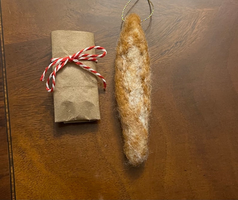 May include: A brown felt ornament shaped like a baguette, hanging from a gold string. The ornament is on a brown wooden surface.