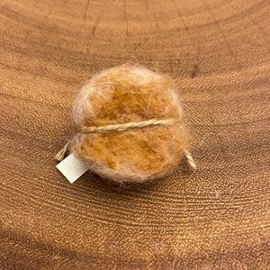 May include: A small, round ball of light brown yarn, tightly wrapped with a natural twine. A small, blank white tag is attached. The yarn ball sits on a textured, brown wooden surface.