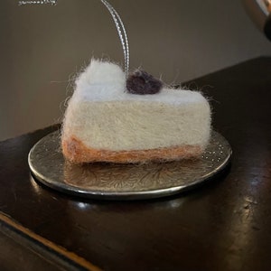 May include: A felt ornament shaped like a slice of cheesecake, with a white fluffy topping and a dark purple berry. The cheesecake sits on a small, silver, decorative plate. A silver string is attached for hanging. The ornament is a dessert-themed decoration.
