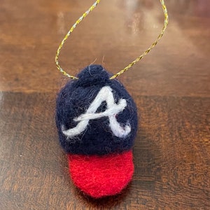 May include: A handmade felt baseball cap ornament. The cap is navy blue with a white "A" and a red brim. A gold string is attached for hanging. This item is perfect for sports fans.