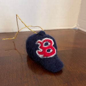 May include: A felt ornament shaped like a baseball cap with a red "B" on the front. The cap is navy blue with a white outline around the "B". The ornament has a gold string for hanging.