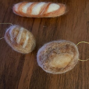 May include: Three felted bread ornaments. The top ornament is a baguette shape with white and orange stripes. The other two ornaments are round, one with a light brown color and the other with a darker brown color. Each ornament has a gold string for hanging.