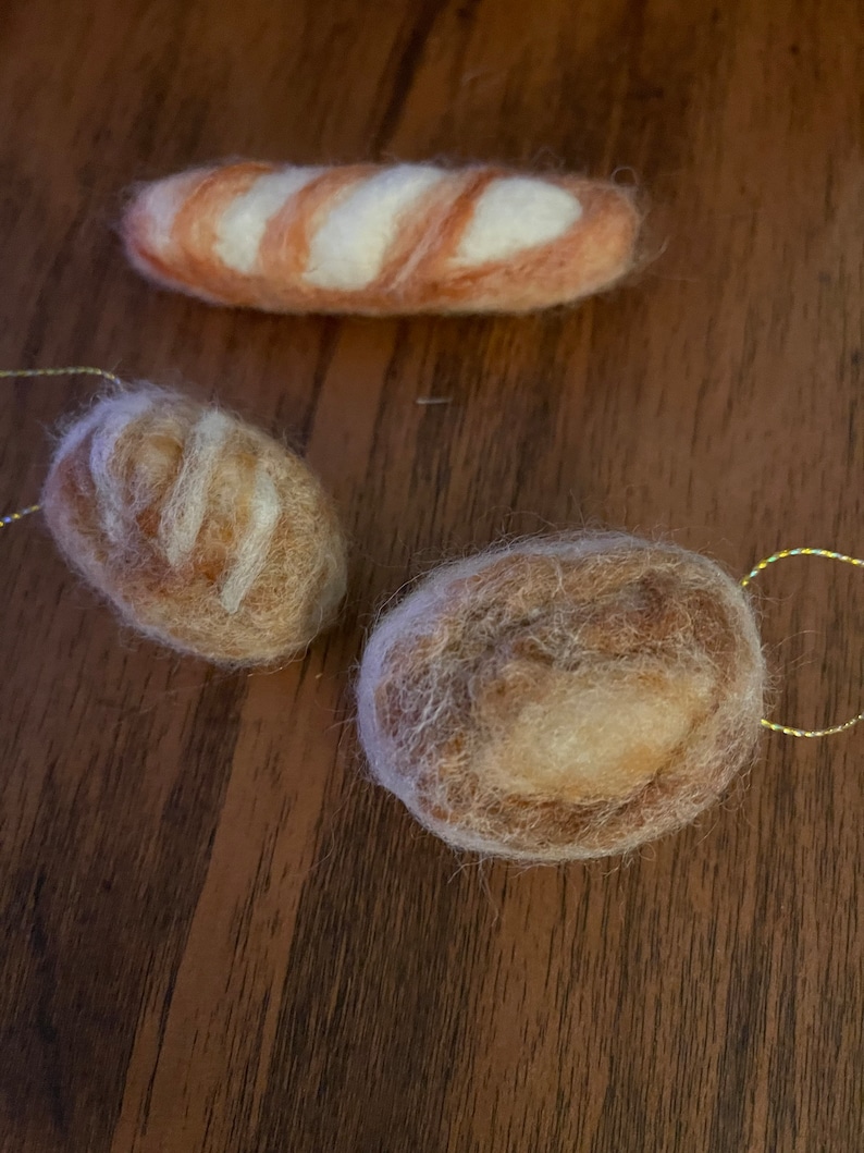 May include: Three felted bread ornaments. The top ornament is a baguette shape with white and orange stripes. The other two ornaments are round, one with a light brown color and the other with a darker brown color. Each ornament has a gold string for hanging.