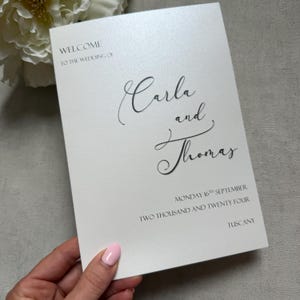 May include: A rectangular white wedding invitation card with a subtle shimmer finish. The text reads: WELCOME TO THE WEDDING OF Carla and Thomas MONDAY 16TH SEPTEMBER TWO THOUSAND AND TWENTY FOUR TUSCANY. The names are in a black cursive script.