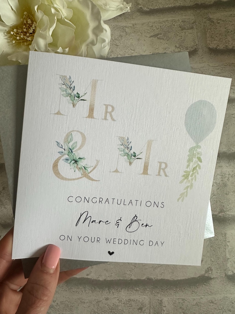 Puede incluir: Una tarjeta de boda blanca con las palabras "Mr & Mr" en letras doradas, adornada con elementos florales de acuarela. La tarjeta dice "Felicidades Marc & Ben en el d&iacute;a de su boda" con un peque&ntilde;o coraz&oacute;n.