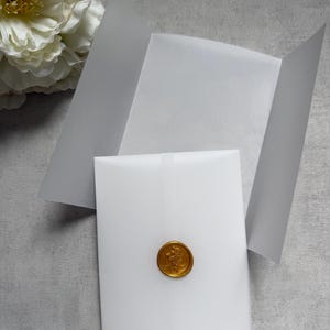 May include: A white envelope sealed with a gold wax seal featuring a floral design. The envelope is partially open, revealing a blank interior. A large white flower is visible in the background, set against a gray surface.