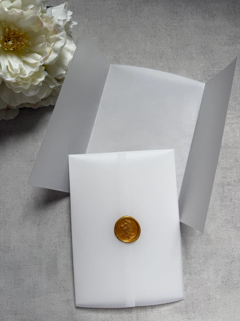May include: A white envelope sealed with a gold wax seal featuring a floral design. The envelope is partially open, revealing a blank interior. A large white flower is visible in the background, set against a gray surface.