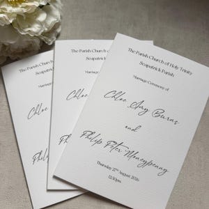 May include: Three white rectangular wedding ceremony programmes with black text. The text reads The Parish Church of Holy Trinity Seapatrick Parish, Marriage Ceremony of Chloe Amy Burns and Philip Peter Moneypenny, Thursday 27th August 2026, 12:30pm.