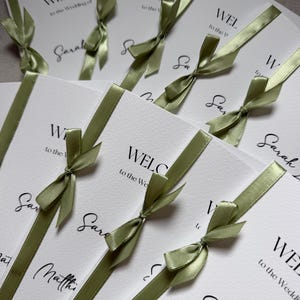 May include: A collection of white wedding programmes featuring black printed text that reads WELCOME to the Wedding of, followed by names like Sarah and Matthew. Each programme is accented with a sage green satin ribbon tied in a small bow.