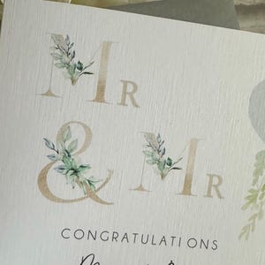 Puede incluir: Tarjeta de felicitaci&oacute;n de boda con la inscripci&oacute;n dorada "Mr & Mr", adornada con motivos florales en acuarela. La tarjeta incluye las palabras "Enhorabuena" y los nombres "Marc & Ben" en cursiva elegante.