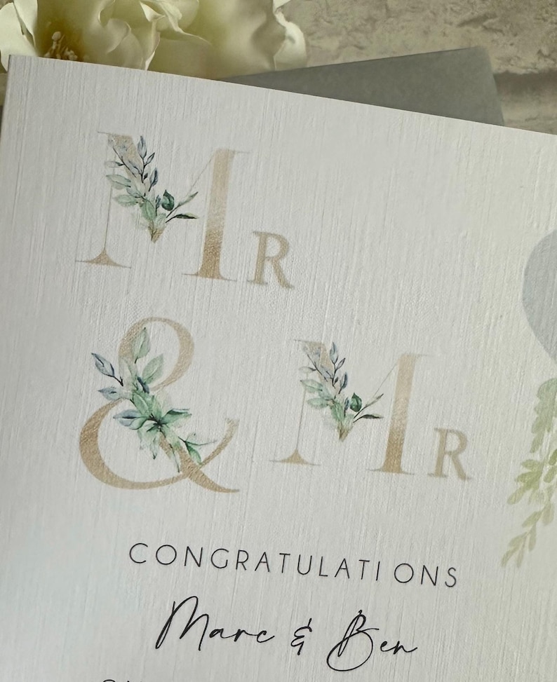 Puede incluir: Tarjeta de felicitaci&oacute;n de boda con la inscripci&oacute;n dorada "Mr & Mr", adornada con motivos florales en acuarela. La tarjeta incluye las palabras "Enhorabuena" y los nombres "Marc & Ben" en cursiva elegante.