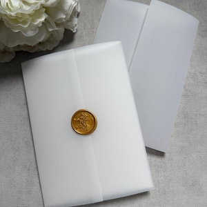 May include: White vellum envelopes sealed with a gold wax stamp. The stamp features a floral design. A large white flower is in the background. The envelopes are on a gray surface.