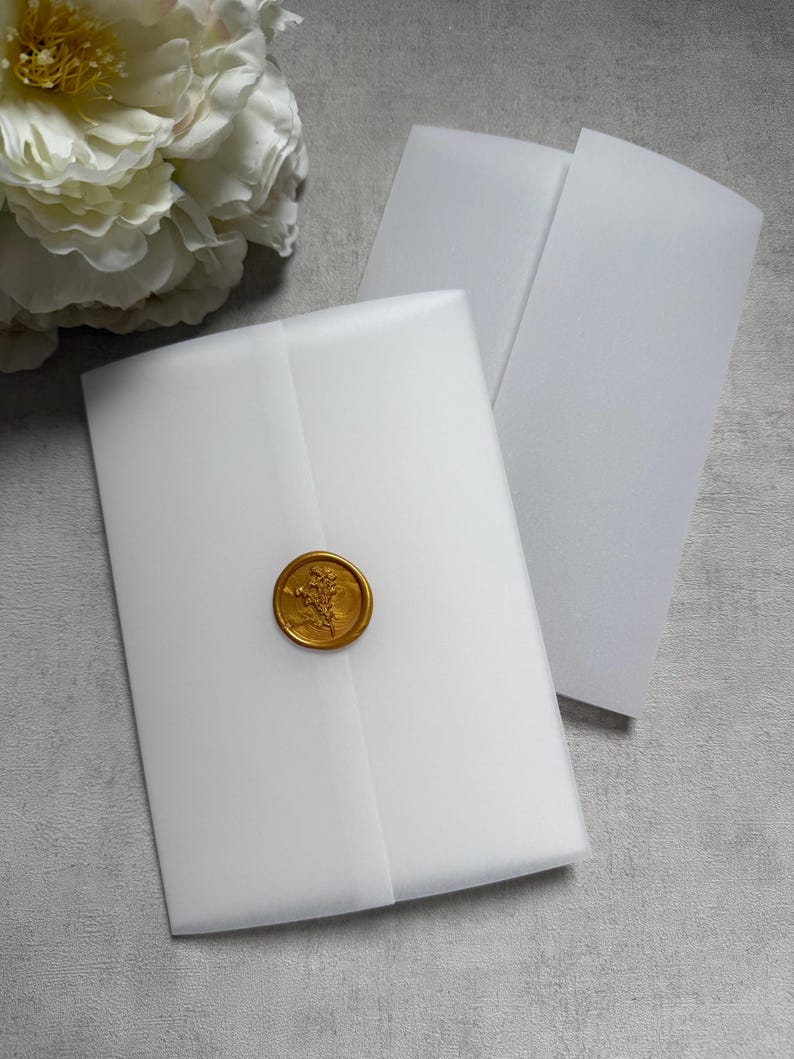 May include: White vellum envelopes sealed with a gold wax stamp. The stamp features a floral design. A large white flower is in the background. The envelopes are on a gray surface.