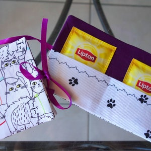 Könnte beinhalten: Zwei Stoffgeschenkkisten, eine mit einem violetten Deckel und die andere mit einem weißen Deckel mit einem schwarz-weißen Katzenmuster. Die violette Kiste enthält zwei gelbe Teebeutel mit dem Text "Lipton" darauf. Die weiße Kiste ist mit einem violetten Band gebunden.