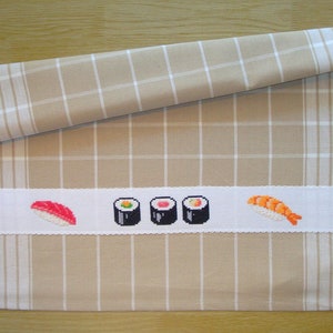 Puede incluir: Paño de cocina a cuadros beige y blanco con una banda bordada blanca con diseños de sushi bordados a punto de cruz. El sushi incluye nigiri y maki. La toalla está enrollada en la parte superior.