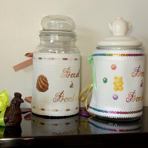 Könnte beinhalten: Zwei Glasbehälter mit dekorativen Stoffhüllen. Einer hat einen Deckel aus klarem Glas, der andere einen weißen Deckel in Teekannenform. Die Stoffhüllen zeigen Kreuzstichmuster, darunter ein Ei und Süßigkeiten, mit dem Schriftzug "Local à Bonbons".