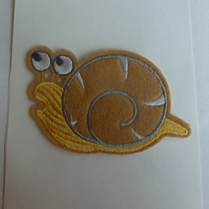 May include: A brown felt snail with a yellow shell and large white eyes. The snail is embroidered with white and gray thread.