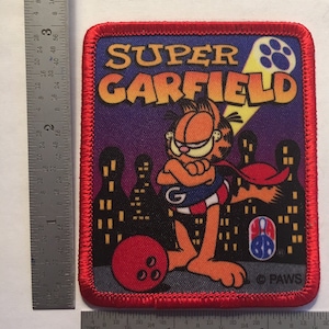 Pode incluir: Um traje de super-herói vermelho, branco e azul com uma capa vermelha e uma grande letra "G" no peito. O personagem é um gato laranja de desenho animado com um nariz vermelho e uma barriga branca. O personagem está segurando uma bola de boliche em uma mão e está em pé na frente de uma paisagem urbana. O texto "SUPER GARFIELD" está no topo da imagem. O texto "PAWS" está na parte inferior da imagem.