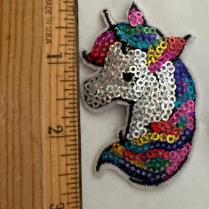 Puede incluir: Un parche con forma de unicornio cubierto de lentejuelas multicolores, incluyendo plata, rosa, azul y amarillo, colocado junto a una regla de madera que muestra una altura de aproximadamente 7,6 cm. El parche tiene un borde blanco y un contorno negro.