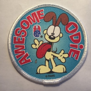 May include: A round, blue and white patch with a cartoon dog named "Odie" from the comic strip "Garfield". The text "AWESOME ODIE" is written in red around the edge of the patch. The dog is yellow with brown ears and a red tongue sticking out. The dog is holding a bowling ball with the number "88" on it.