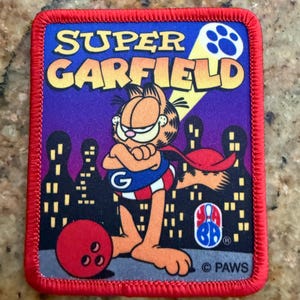 Puede incluir: Un parche rectangular bordado con borde rojo que muestra a Garfield vestido de superhéroe con capa roja y camiseta azul con una G. El texto SUPER GARFIELD aparece sobre una silueta urbana, una bola de bolos roja y un logotipo de YABA.