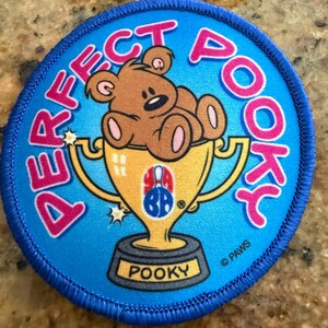 Puede incluir: Un parche bordado circular con fondo azul brillante. Presenta un oso de peluche marrón sentado en una copa de trofeo dorada. El texto "PERFECT POOKY" está bordado en rosa alrededor del oso. La base del trofeo dice "POOKY" y tiene un pequeño logo.