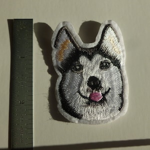 May include: Embroidered patch of a husky dog's face with a pink tongue sticking out. The dog has black and white fur and brown eyes.