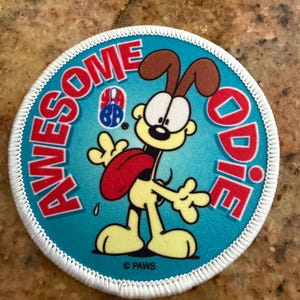 Puede incluir: Un parche bordado circular con un borde blanco sobre un fondo azul brillante. Presenta un perro de dibujos animados con pelaje amarillo y orejas marrones, sacando la lengua roja. El texto AWESOME ODIE está impreso en rojo, con un pequeño logo YABA.