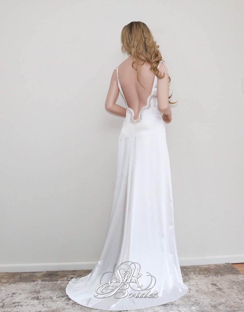 May include: A white satin wedding dress with a low back and a lace trim. The dress has a flowing skirt and a simple, elegant design.
