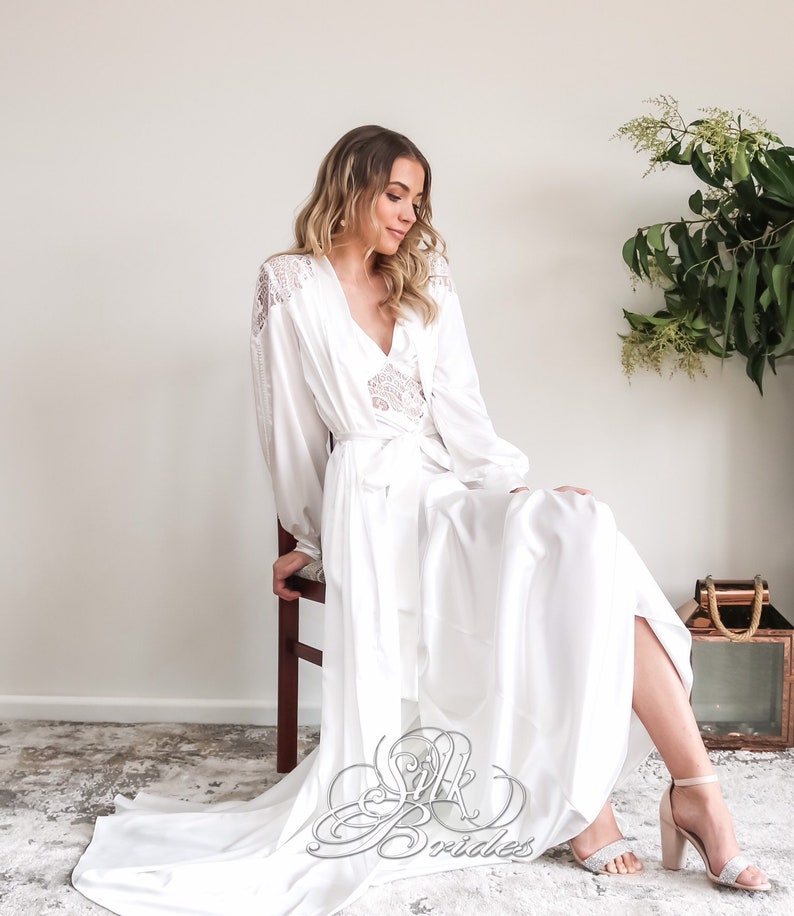 May include: A white satin robe with long sleeves and a lace trim. The robe has a sash tie at the waist and a flowing skirt. The model is wearing the robe and sitting on a chair.
